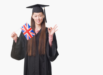 Young Chinese woman wearing graduate uniform holding United kingdom flag doing ok sign with fingers, excellent symbol