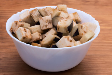 Eggplant, diced, stacked in white porcelain plate on a wooden background.