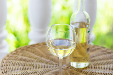 Glass of white dry wine and bottle on wicker table on garden terrace of villa or mansion. Authentic lifestyle image. Relaxation indulgence gourmet. Poster with copy space