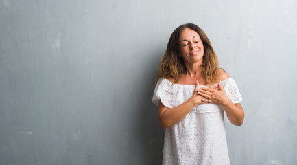 Middle age hispanic woman standing over grey grunge wall smiling with hands on chest with closed eyes and grateful gesture on face. Health concept.