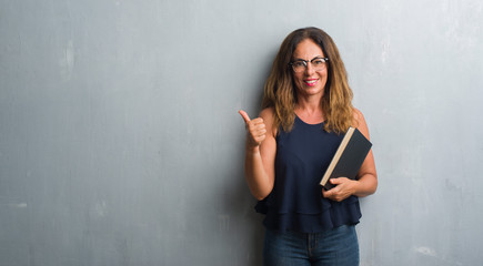 Middle age hispanic woman standing over grey grunge wall holding a book happy with big smile doing...