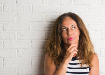 Middle age hispanic woman over white brick wall serious face thinking about question, very confused...