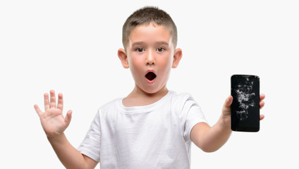 Dark haired little child holding broken smartphone scared in shock with a surprise face, afraid and...