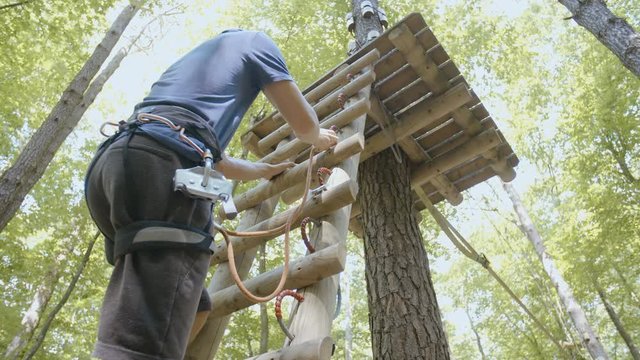 Person climb the ladder while attaching safety clips in slow motion 4K. Low angle of a young climber in focus with safety equipment rising up the wooden ladder to a tree platform in the forest.