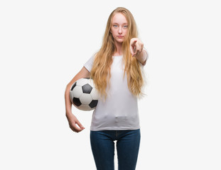 Blonde teenager woman holding soccer football ball pointing with finger to the camera and to you, hand sign, positive and confident gesture from the front