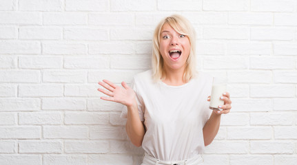 Adult caucasian woman over white brick wall drinking glass of milk very happy and excited, winner expression celebrating victory screaming with big smile and raised hands