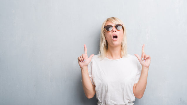 Adult caucasian woman over grunge grey wall wearing sunglasses amazed and surprised looking up and pointing with fingers and raised arms.