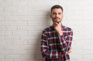 Young adult man standing over white brick wall looking confident at the camera with smile with crossed arms and hand raised on chin. Thinking positive.