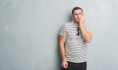 Young caucasian man over grey grunge wall looking stressed and nervous with hands on mouth biting nails. Anxiety problem.