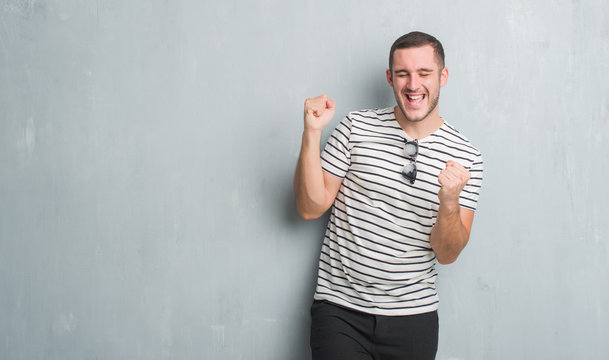 Young caucasian man over grey grunge wall very happy and excited doing winner gesture with arms raised, smiling and screaming for success. Celebration concept.