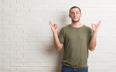 Young caucasian man standing over white brick wall relax and smiling with eyes closed doing meditation gesture with fingers. Yoga concept.