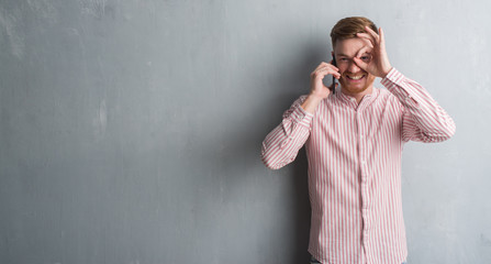 Young redhead man over grey grunge wall talking on the phone with happy face smiling doing ok sign with hand on eye looking through fingers