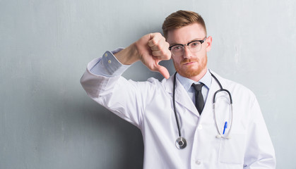 Young redhead doctor man over grey grunge wall with angry face, negative sign showing dislike with...