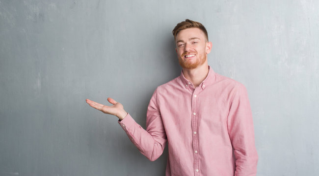 Young redhead man over grey grunge wall wearing pink shirt smiling cheerful presenting and pointing with palm of hand looking at the camera.