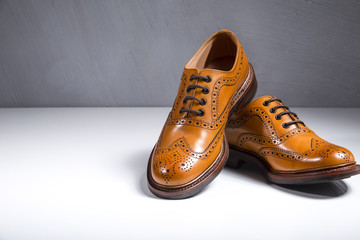 Fashionable Luxury Male Full Broggued Tan Leather Oxfords Shoes Placed Over White Surface. Against Gray Wall Background.
