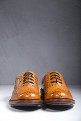 Luxury Male Full Broggued Tan Leather Oxfords Shoes Placed Over White Surface. Against Gray Wall.