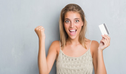 Beautiful young woman over grunge grey wall holding credit card screaming proud and celebrating...