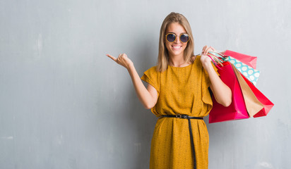 Beautiful young woman over grunge grey wall holding shopping bags on sales very happy pointing with...