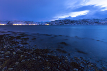 Sunset at the lakeside with rocks of a fjord during low tide in a snowy winter landscape.