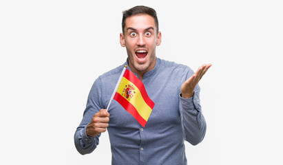 Handsome young man holding a flag of Spain very happy and excited, winner expression celebrating victory screaming with big smile and raised hands