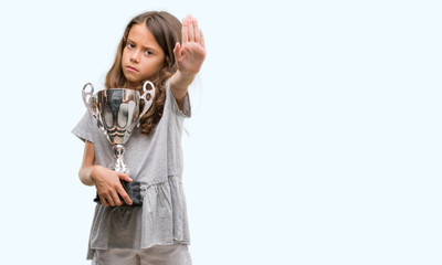 Brunette hispanic girl holding a trophy with open hand doing stop sign with serious and confident expression, defense gesture