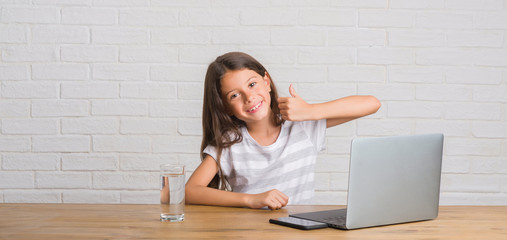 Young hispanic kid sitting on the table using computer laptop happy with big smile doing ok sign,...