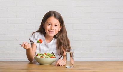 Young hispanic kid sitting on the table eating healthy salad with a happy face standing and smiling...