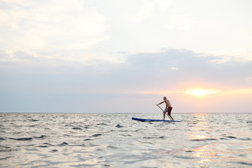 A man stands on a SUP board against the background of the sea and the sunset.