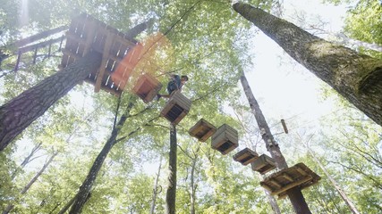 Person walk over boxes hanging between trees in adventure park 4K. Low angle wide shot handheld slide of a person in focus carefully walking towards another tree platform. Sunflares shine in the frame