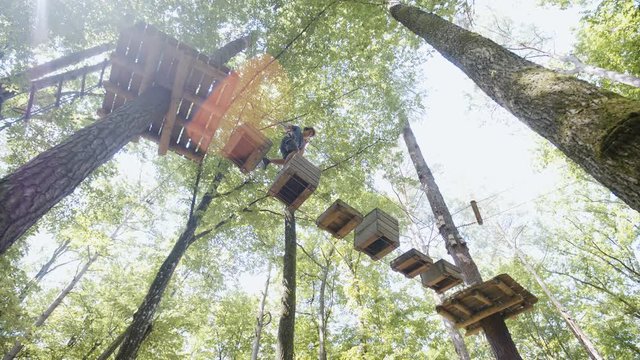 Person walk over boxes hanging between trees in adventure park 4K. Low angle wide shot handheld slide of a person in focus carefully walking towards another tree platform. Sunflares shine in the frame