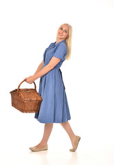 full length portrait of blonde girl wearing blue dress holding a wicker picnic basket. standing pose. isolated on white  studio background.