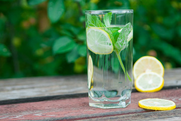 Cold lemonade with lemon, mint and berries in a transparent glass with facets. On an old wooden table. In the background, the green foliage is a soft blurred focus.