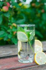 Water with lemon and mint in a transparent glass with facets. On an old wooden table. In the background, the green foliage is a soft blurred focus.
