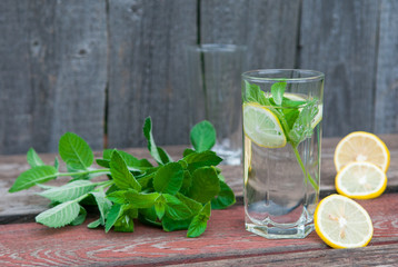 Water with lemon and mint in a transparent glass with facets. On an old wooden table. In the background, the green foliage is a soft blurred focus.