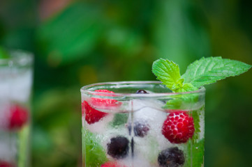 Cold lemonade with lemon, mint and berries in a transparent glass with facets. On an old wooden table. In the background, the green foliage is a soft blurred focus.