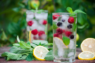 Cold lemonade with lemon, mint and berries in a transparent glass with facets. On an old wooden table. In the background, the green foliage is a soft blurred focus.