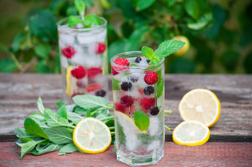 Cold lemonade with lemon, mint and berries in a transparent glass with facets. On an old wooden table. In the background, the green foliage is a soft blurred focus.