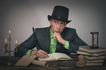 Pensive man in bowler hat and suit is sitting by table and holding a feather in pen in his hand....