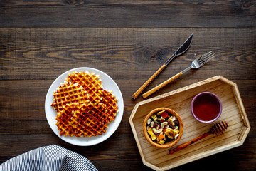Round belgian waffles for breakfast. Breakfast in bed. Waffles on plate. Honey and dried fruits in tray, knife and fork, tablecloth on dark wooden background top view copy space