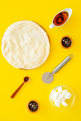 Raw rolled out pizza dough near cheese mozzarella, olive oil, spices, knife for pizza on yellow background top view mockup