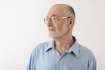 Fototapeta premium Sideways picture of serious handsome sixty year old senior man with gray beard and bald head posing in studio wearing eyeglasses and blue shirt, looking away with pensive thoughtful expression