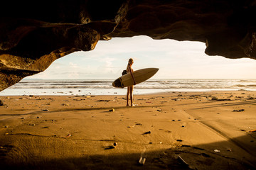 Young surfer woman with surf board pose in cave at beach. Garbage and plastic on sand. Concept of ecological problem and disaster. People water sport adventure camp and extreme swim on summer vacation