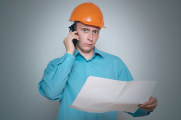 Builder construction worker with mobile phone is holding in hand a plan draft document isolated on gray background.