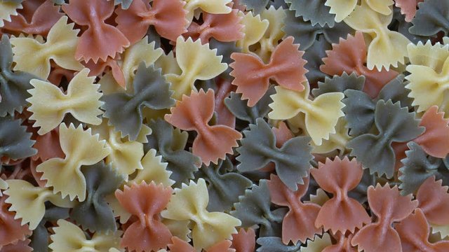Multicolored pasta background, rotating, top view. Rotation of colored farfalle pasta. Heap of uncooked pasta. Italian food close up. Delicious italian farfalle tricolor pasta, food background