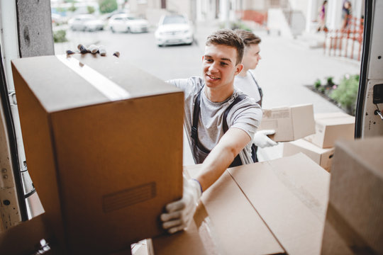 A young handsome smiling mover wearing uniform is reaching for the box while unloading the van full of boxes. House move, mover service.