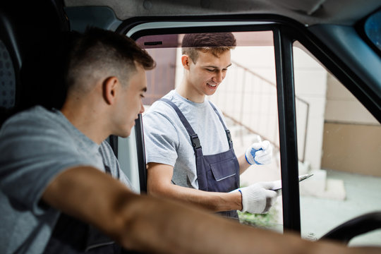 A Young Handsome Guy Wearing Uniform Is Looking Out Of The Car Window. Another Worker Wearing Uniform Is Holding The Clipboard. House Move, Mover Service.