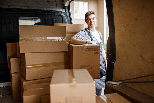 A young handsome smiling worker wearing uniform is standing next to the van full of boxes. House move, mover service.