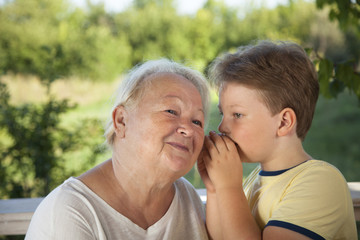 Boy whispering to grandma in summer outdoors garden, grandkid and grandmother lalking