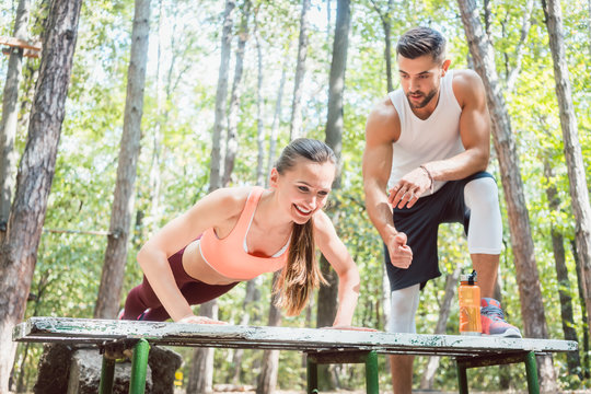 Sporty woman doing push-up in an outdoor gym, her boyfriend is watching her 