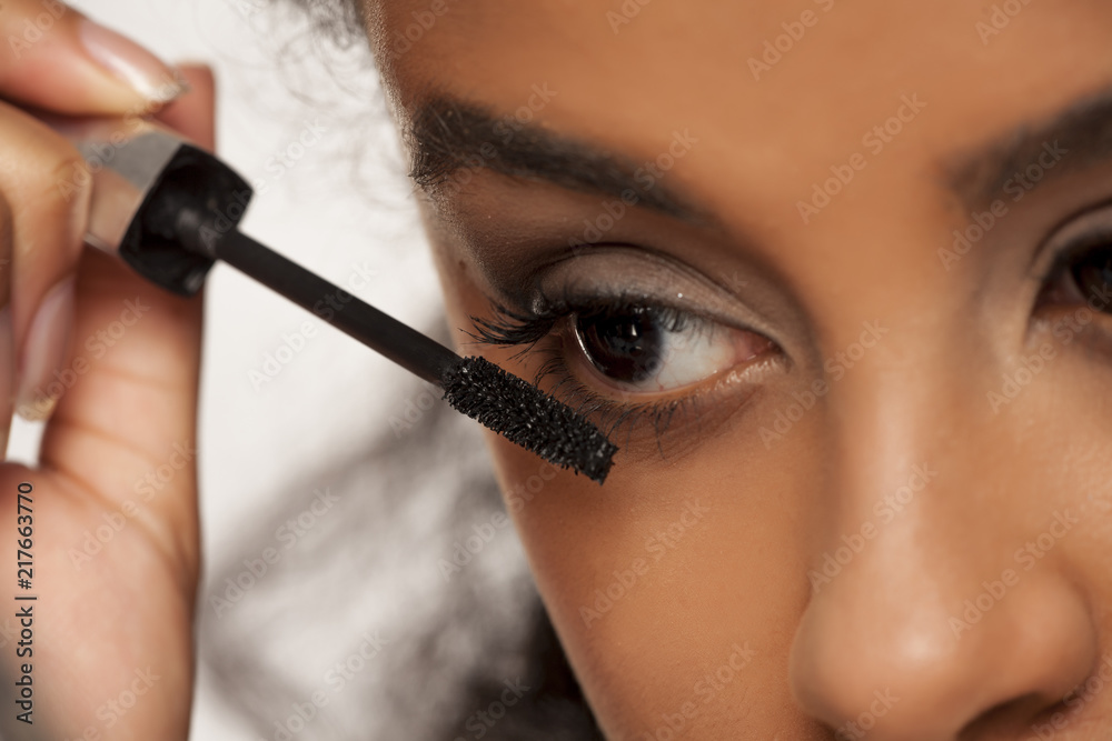 Wall mural portrait of a young dark-skinned woman applying mascara on a white background